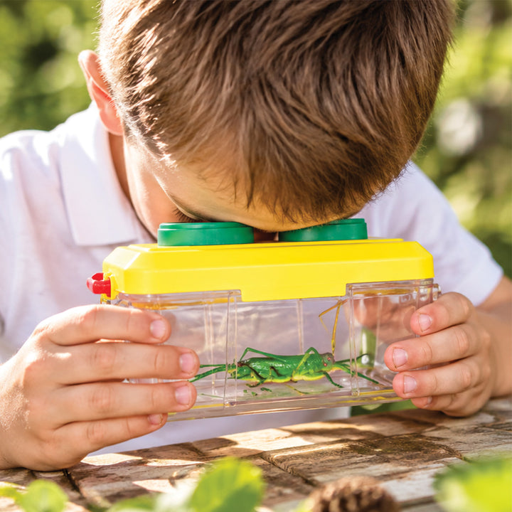 A child outdoors looking through a clear plastic insect viewing box with a yellow top and green eyepieces, holding a green grasshopper inside.